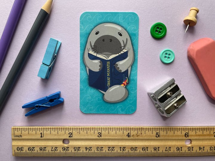 A bookmark of a grey manatee reading a book called "The Great Manatee" on the front and the text "Read Books. Love Manatees." on the back is shown on a purple background surrounded by small objects.