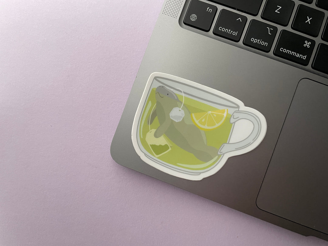 A sticker of a cup of green tea with a tea bag and slice of lemon with a grey manatee swimming in it is shown on a silver laptop.