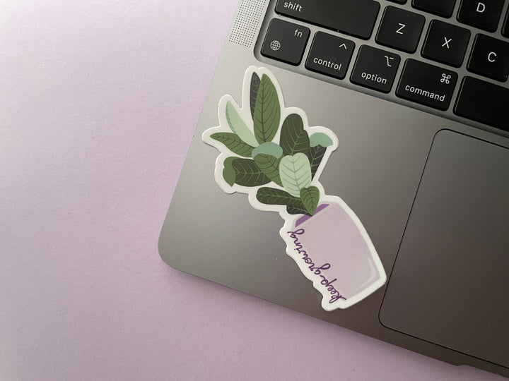 A sticker of a cartoon large-leafed green plant in a purple planter with the text "keep growing" in script up the side of the planter is shown on a silver laptop.