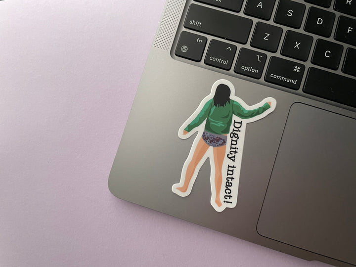 A sticker of a cartoon comedian Aisling Bea dancing away not wearing trousers from the TV show Taskmaster next to the text "Dignity Intact" is shown on a silver laptop.