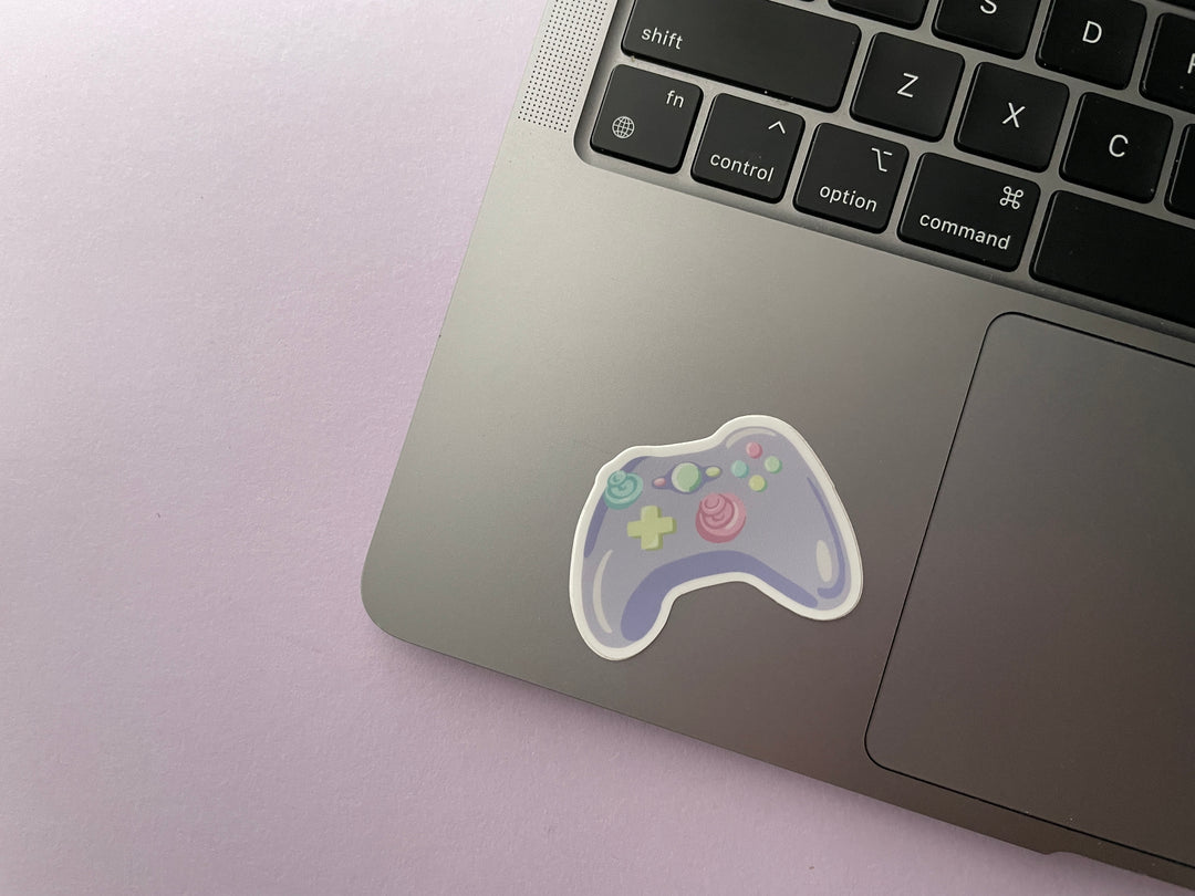 A sticker of a cartoon purple game controller with multi color buttons is shown on a silver laptop.