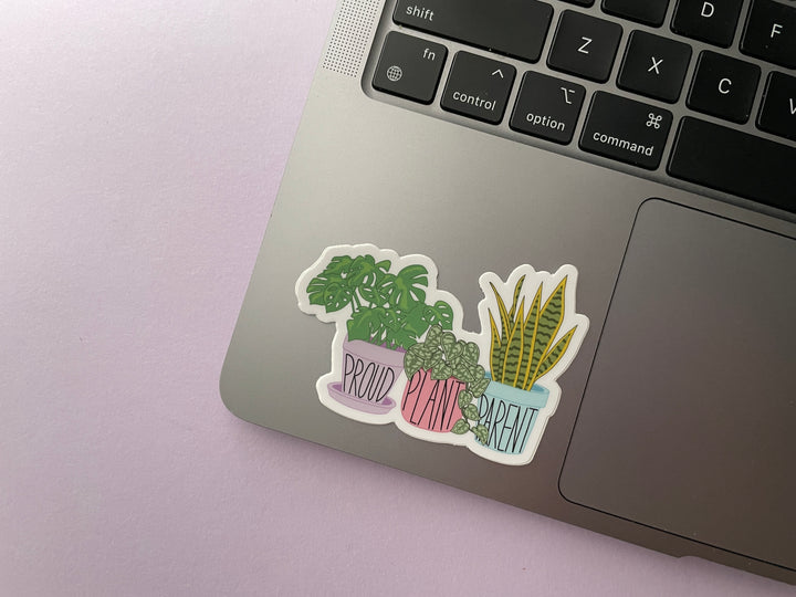 A sticker of a cartoon of three house plants in purple, pink, and blue planters with the text "proud plant parent" is shown on a silver laptop.