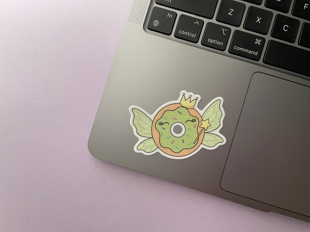 A sticker of a cartoon donut with green frosting with green fairy wings and a starry magic wand and crown is shown on a silver laptop.