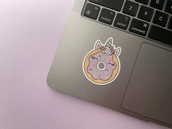 A sticker of a cartoon donut with pink frosting with horse ears and a green and blue striped unicorn horn is shown on a silver laptop.