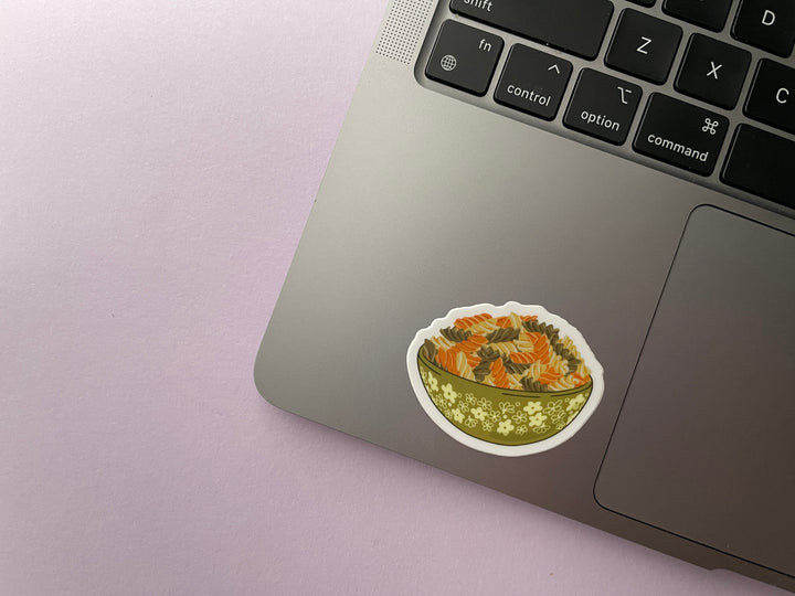 A sticker of a green bowl of orange, green, and cream colored spiraled rotini noodles is shown on a silver laptop.