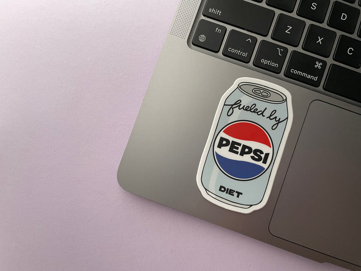 A sticker of a cartoon of a light blue can of Diet Pepsi with the text "fueled by" in the top corner is shown on a silver laptop.