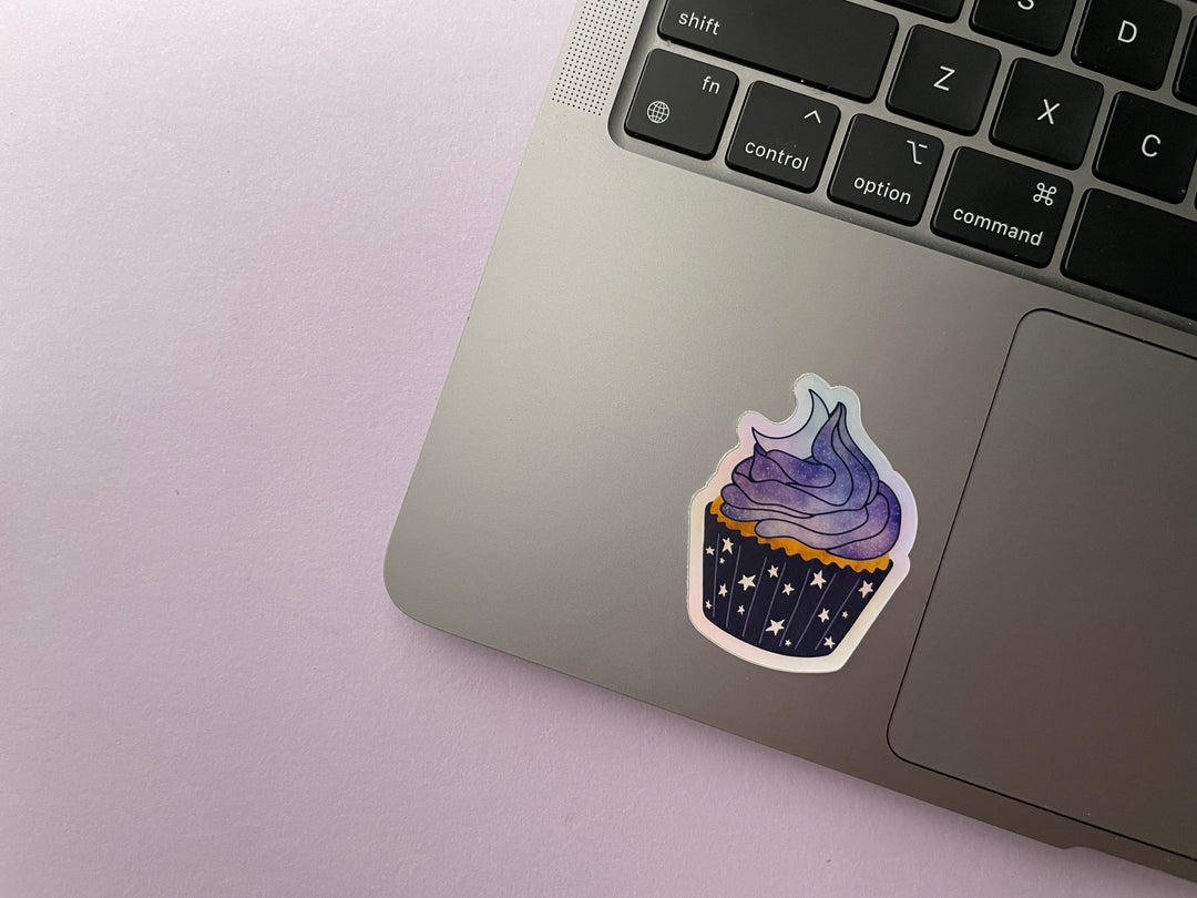 A holographic sticker of a cartoon of a cupcake with starry paper, purple and blue stardust frosting, and top with a crescent moon is shown on a silver laptop.