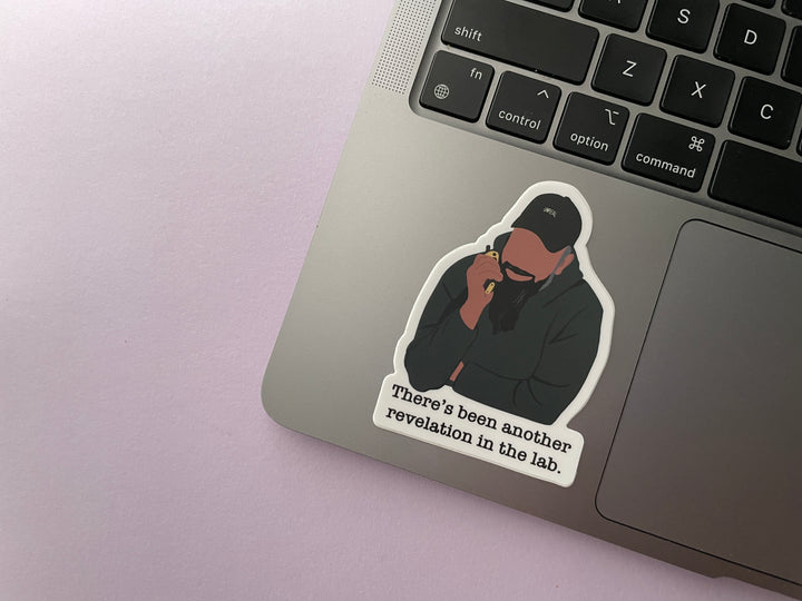 A sticker of a cartoon of comedian Guz Khan holding a walkie talkie with the text "there's been another revelation in the lab" belowis shown on a silver laptop.