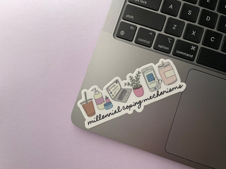 A sticker of a cartoon of a series of items including a candle, laptop, houseplant, iced coffee, lotion, and phone above the words "millennial coping mechanisms" is shown on a silver laptop.