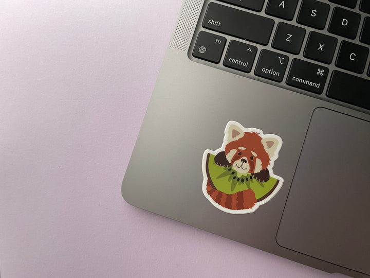 A sticker of a cartoon red panda holding a large slice of kiwi fruit is shown on a silver laptop.