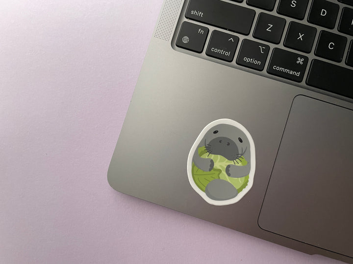 A sticker of a cartoon grey manatee with flippers wrapped around a head of green lettuce is shown on a silver laptop.