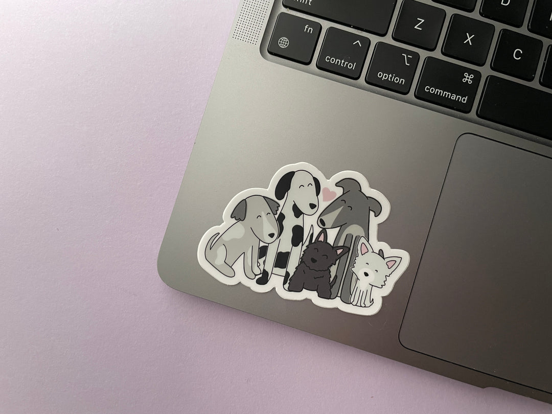 A sticker of a cartoon drawing of five dogs of different sizes in black and white sitting together with a pink heart above them is shown on a silver laptop.