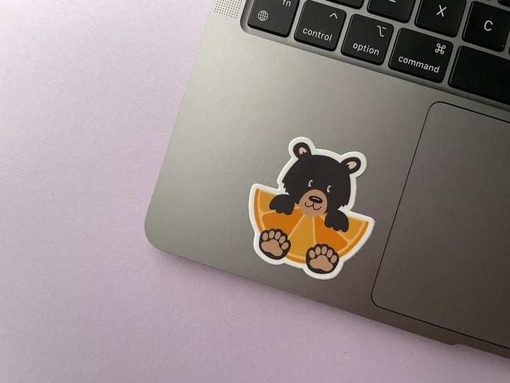 A sticker of a cartoon black bear holding a large orange fruit slice is shown on a silver laptop.