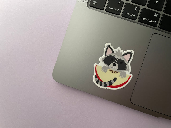 A sticker of a cartoon raccoon holding a large slice of red apple is shown on a silver laptop.