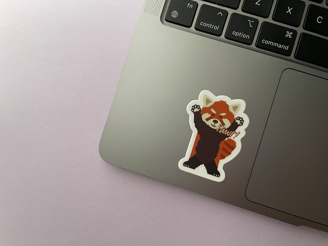 A sticker of a cartoon red panda standing on its hind legs with its arms raised and the text "rawr" next to its mouthis shown on a silver laptop.