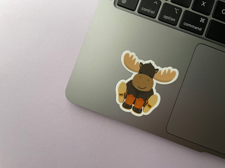 A sticker of a smiling cartoon moose sitting with orange and brown boots on its hooves is shown on a silver laptop.