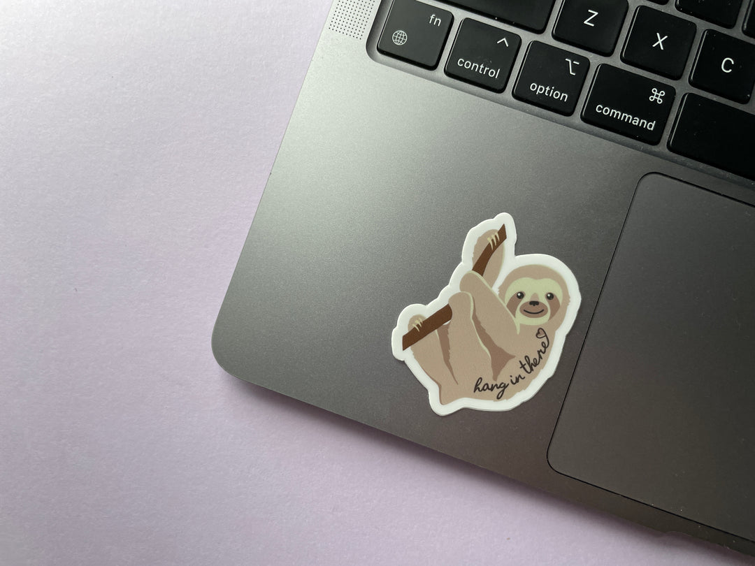 A sticker of a light brown sloth in profile hanging from a brand with the text "hang in there" and a heart over top is shown on a silver laptop.