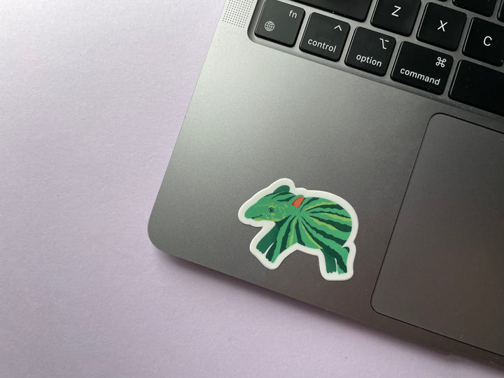 A sticker of a green cartoon tapir with watermelon stripes along its body is shown on a silver laptop.