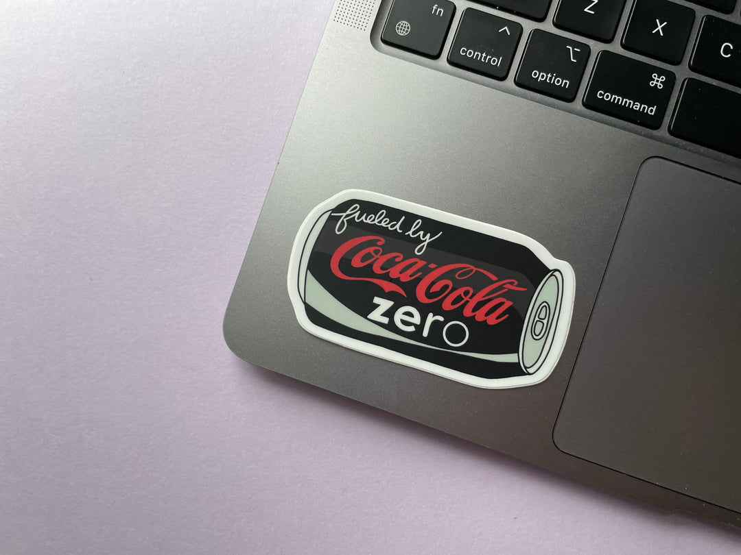 A sticker of a cartoon of a black can of Coke Zero on its side with the text "fueled by" in the top corner is shown on a silver laptop.