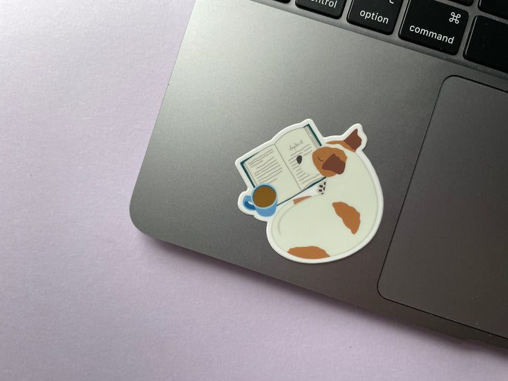 A sticker of a brown and white dog curled up next to an open box and mug of brown liquid is shown on a silver laptop.