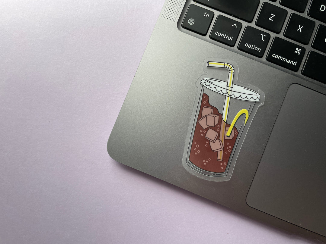 A sticker of a clear fountain drink cup with part of a yellow "M" and a bendy straw three-quarters filled with ice and bubbly brown liquid is shown on a silver laptop.