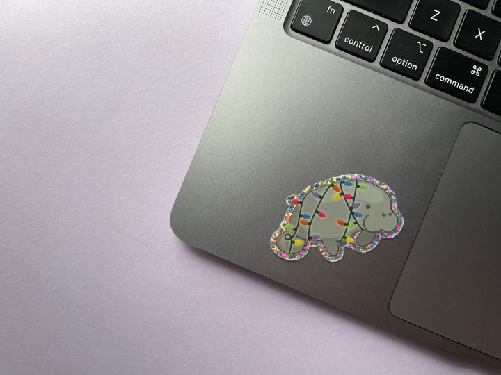 A glittery sticker of a cartoon grey manatee shown in profile wrapped in multi-color holiday lights is shown on a silver laptop.