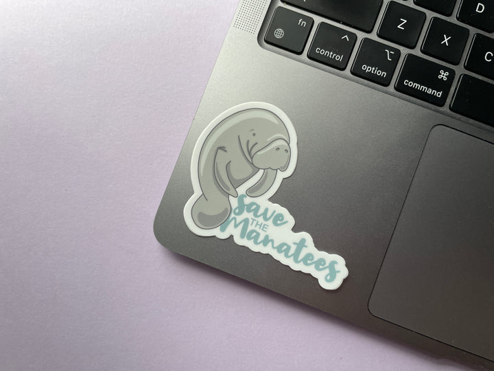 A sticker of a grey cartoon manatee standing of the size next to the light blue text "save the manatees" is shown on a silver laptop.