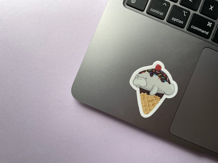 A sticker of a cartoon ice cream cone dipped in chocolate topped with a grey manatee with chocolate sauce, sprinkles, and a cherry is shown on a silver laptop.