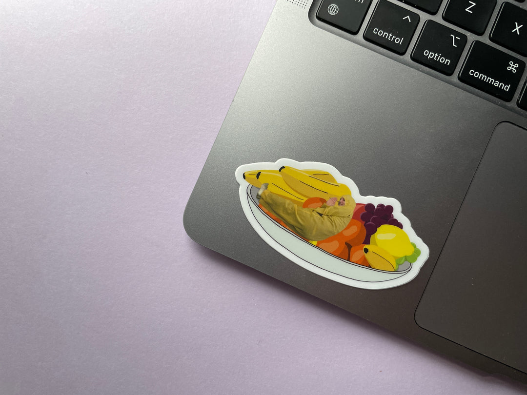 A sticker of a cartoon of comedian Noel Fielding in a yellow jumpsuit disguised in a bowl of fruit from the TV show Taskmaster is shown on a silver laptop.