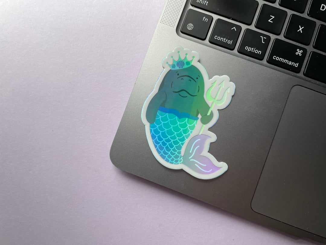 A holographic sticker of a cartoon grey manatee with a multi colored mermaid tail and yellow crown holding a yellow trident is shown on a silver laptop.