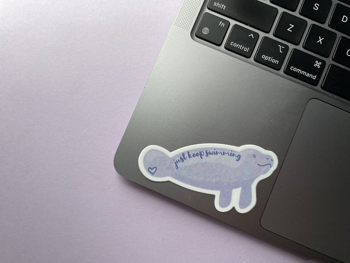 A sticker of a cartoon light purple smiling manatee shown from the side with the text "just keep swimming" over top is shown on a silver laptop.