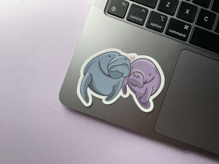 A sticker of a cartoon pair of manatees, one blue and one purple, with their heads together and a heart about them is shown on a silver laptop.