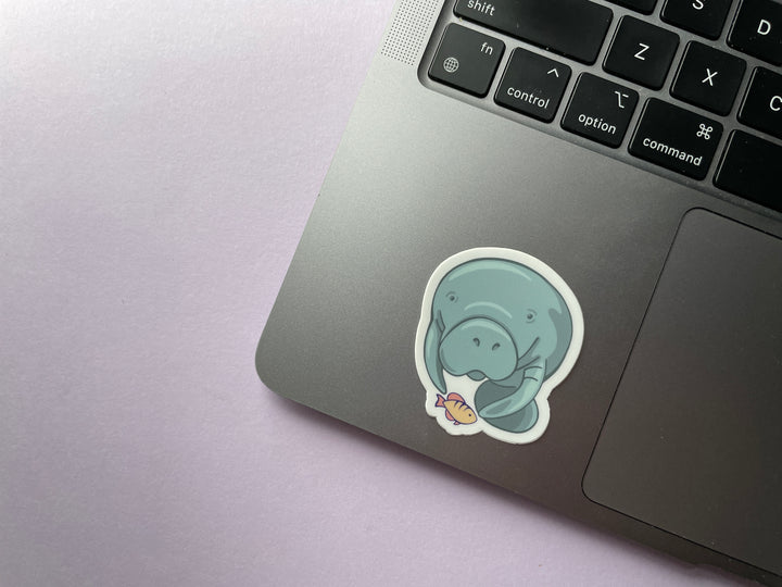 A sticker of a cartoon blue manatee head on with a yellow and orange fish beneath it is shown on a silver laptop.