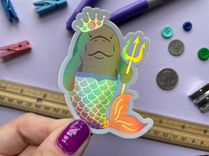 A holographic sticker of a cartoon grey manatee with a multi colored mermaid tail and yellow crown holding a yellow trident is held over a purple background covered in objects.