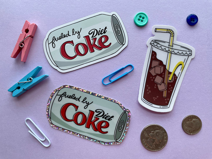 Three items on a purple background surrounded by objects. The items are a Diet Coke magnet, Diet Coke glitter sticker, and a fountain drink with bendy straw sticker.