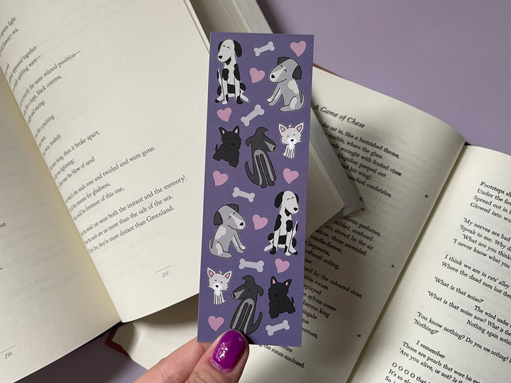 A purple bookmark covered in pink hearts and black and white drawings of cartoon dogs is held above open books and a purple background.