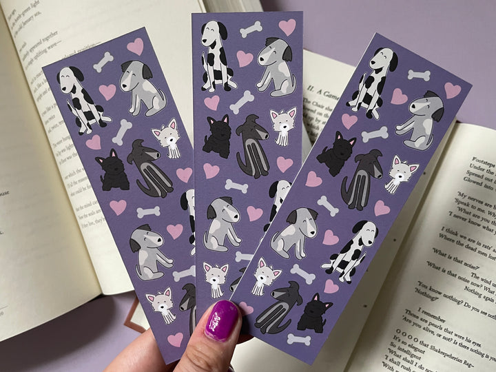 Three purple bookmarks covered in pink hearts and black and white drawings of cartoon dogs is held above open books and a purple background.