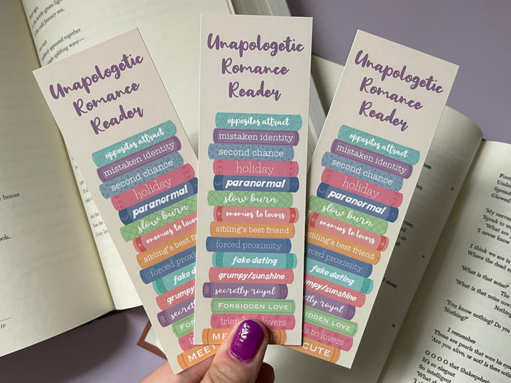 Three tan bookmarks of a stack of multicolored books with different romance tropes along the spines and the text "Unapologetic Romance Reader" along the top are held above books and a purple background.