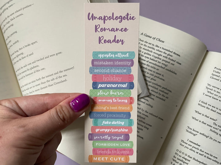 A tan bookmark of a stack of multicolored books with different romance tropes along the spines and the text "Unapologetic Romance Reader" along the top is held above open books and a purple background.