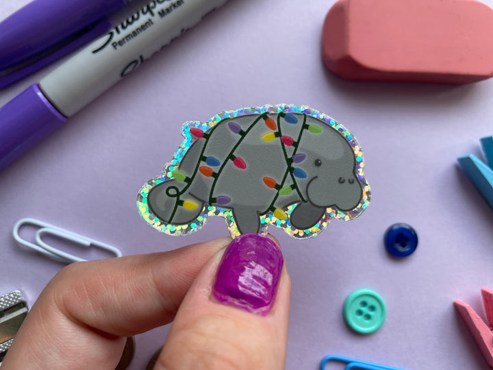 A glittery sticker of a cartoon grey manatee shown in profile wrapped in multi-color holiday lights is shown over a purple background covered with small objects.