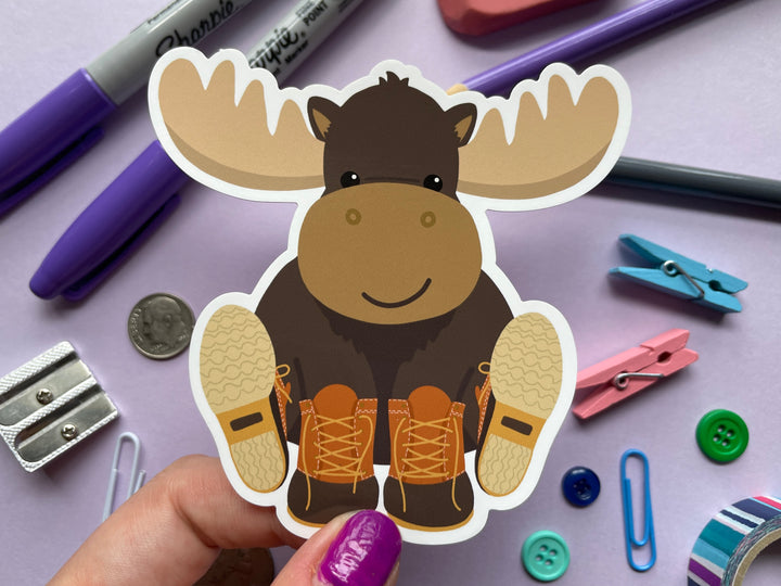 A static cling of a smiling cartoon moose sitting with orange and brown boots on its hooves is held over a purple background covered in objects.