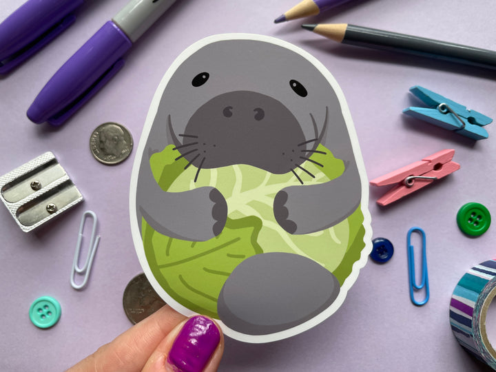 A static cling of a cartoon grey manatee with flippers wrapped around a head of green lettuce is held over a purple background covered in objects.