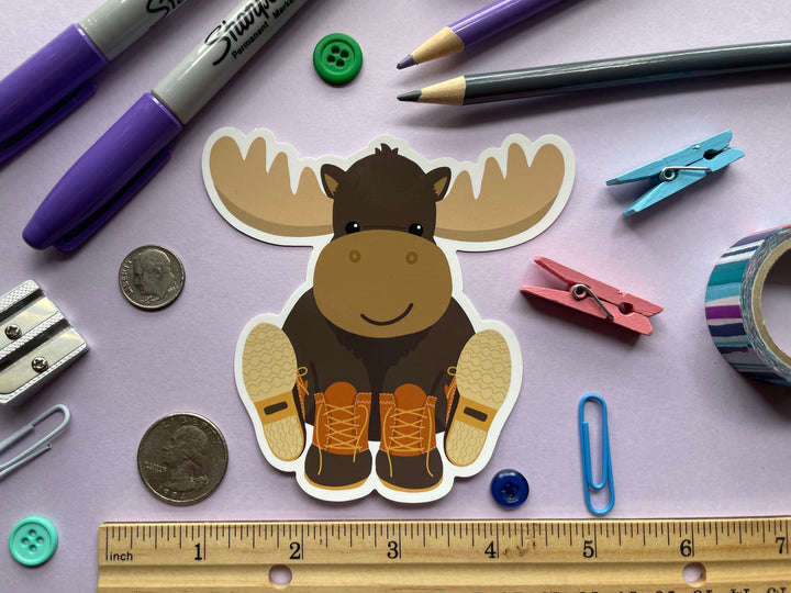 A static cling of a smiling cartoon moose sitting with orange and brown boots on its hooves is shown on a purple background surrounded by small objects.