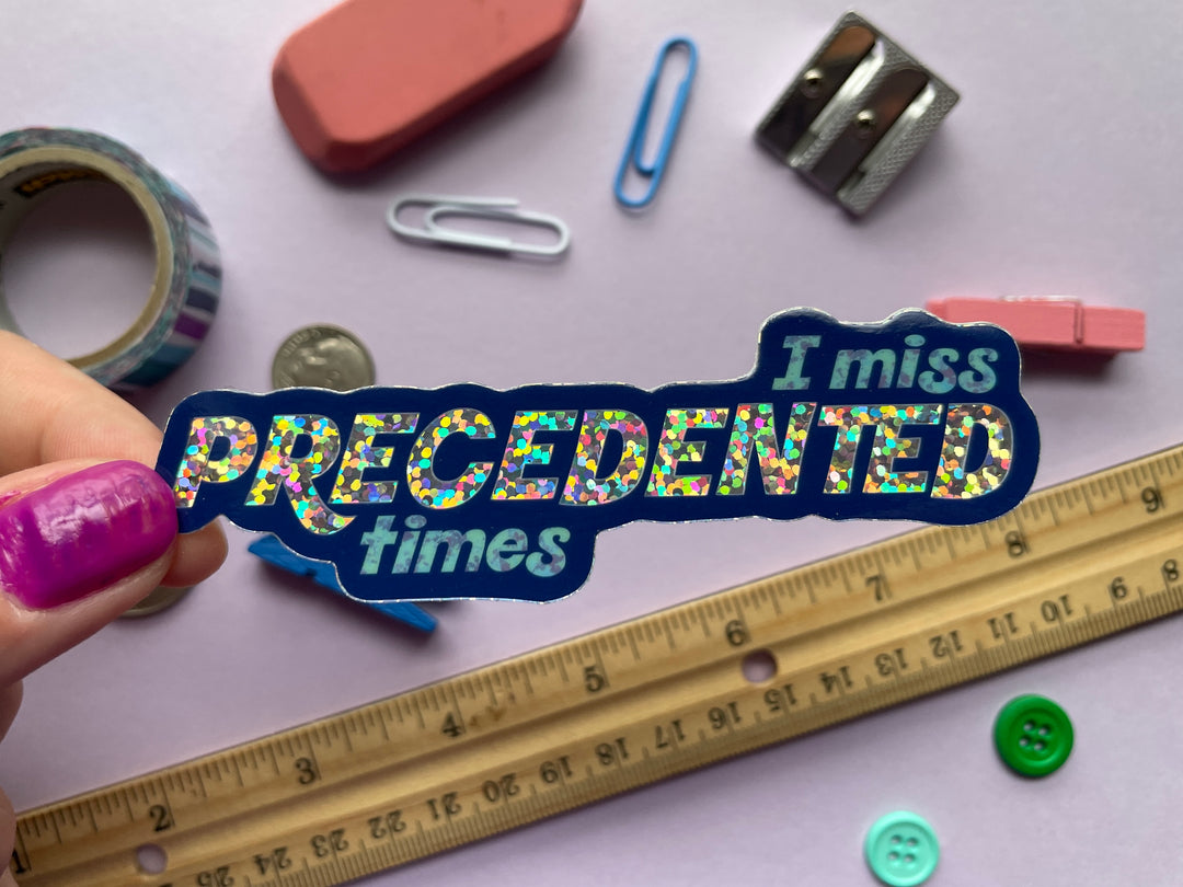 A blue and glittery sticker with block text that says "I miss precedented times" is held above a purple background covered in small objects.