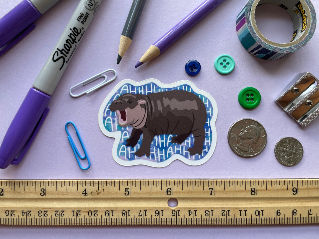 A sticker of a brown baby pygmy hippo with its mouth open against a blue background with the repeating text "AHHHHH" is shown on a purple background surrounded by small objects such as coins, buttons, and a ruler.