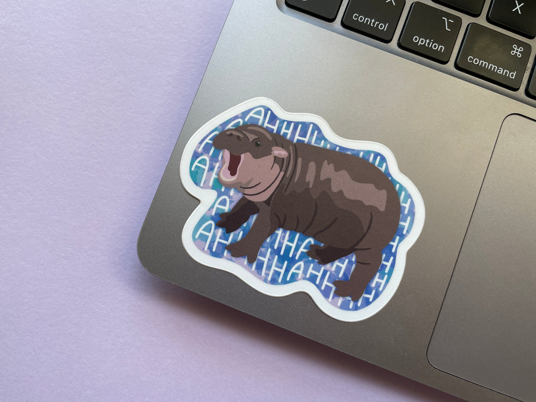 A sticker of a brown baby pygmy hippo with its mouth open against a blue background with the repeating text "AHHHHH" is shown on silver laptop on a purple background.