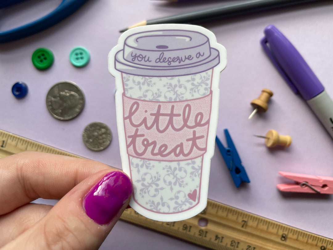 A sticker of a pastel paper coffee cup with a purple lid and a pink coffee sleeve is held above of a purple background. Scripting text on the cup reads "you deserve a little treat".