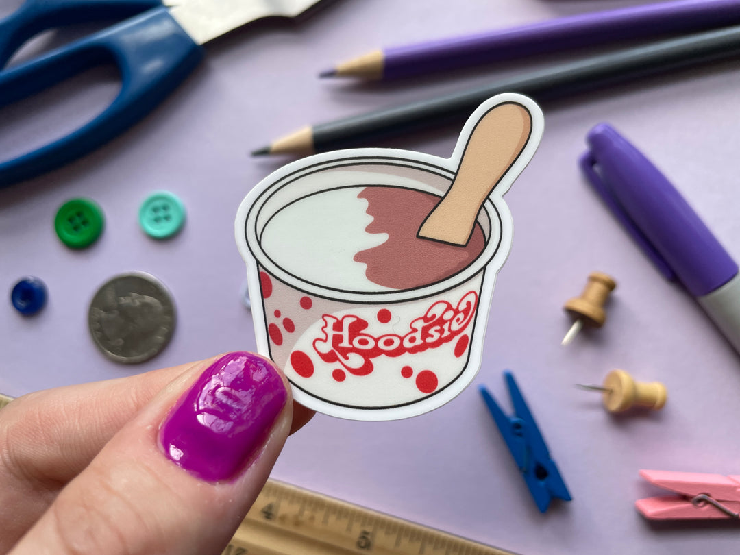 A sticker of a white and red Hoodsie cup with vanilla and chocolate ice cream and a wooden spoon is held over a purple background.