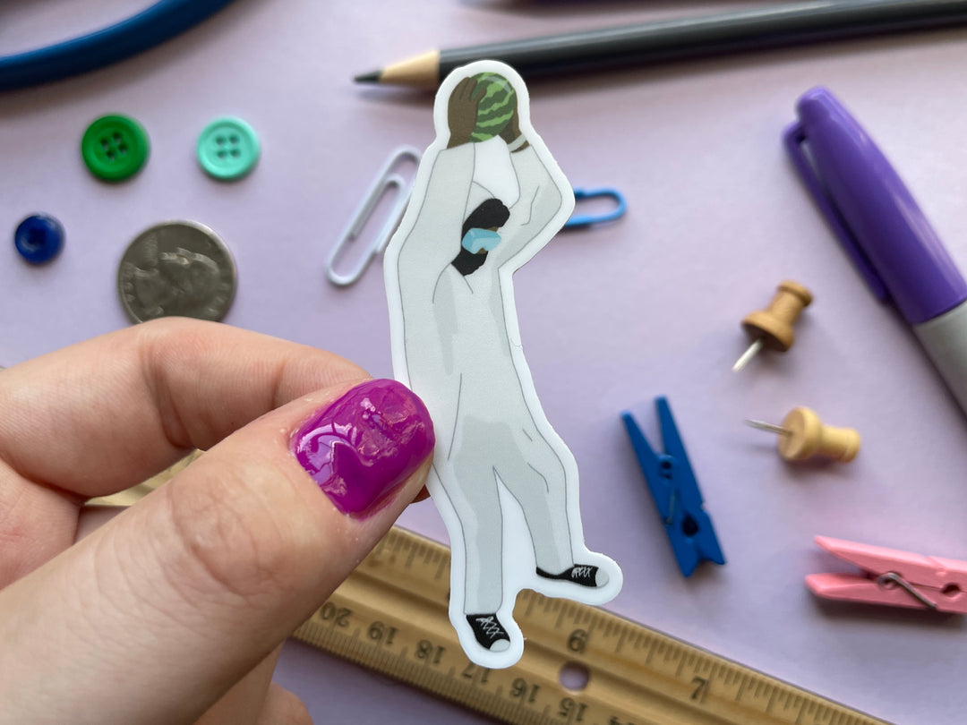 A sticker of comedian Romesh Ranganathan in a white jumpsuit holding a watermelon above his head is shown held over a purple background covered in small objects.