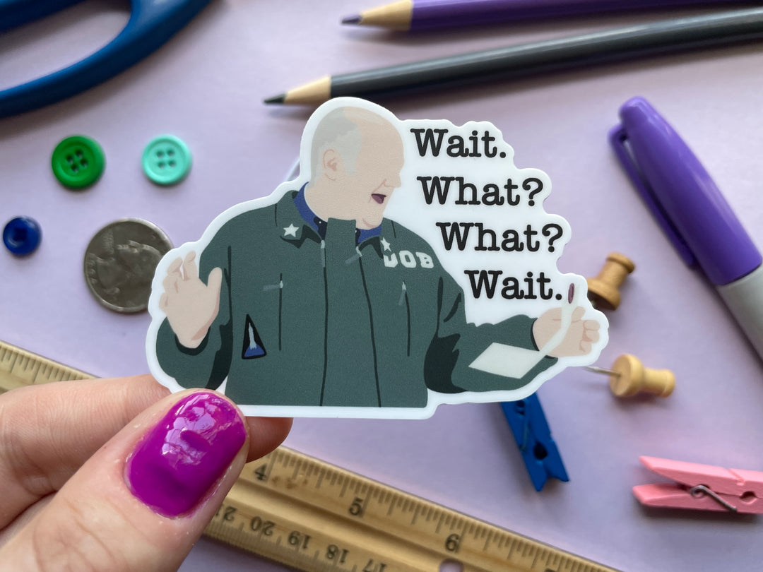 A sticker of a cartoon image of comedian Dara O Briain with his arms up holding a paper from the TV show Taskmaster with the text &quot;Wait. What? What? Wait.&quot; is shown held over a purple background covered in small objects.